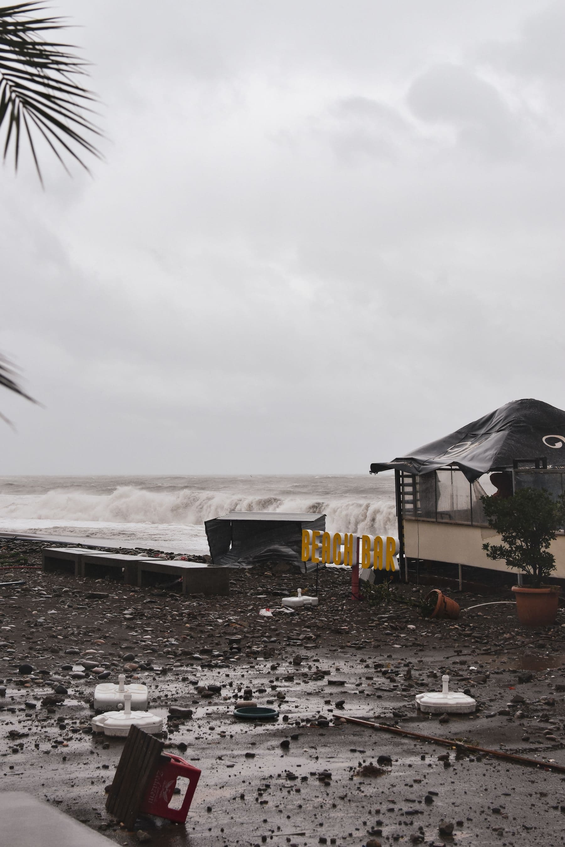 A storm-damaged beachfront bar under a gray sky, with rough surf behind it and debris scattered across the sand.