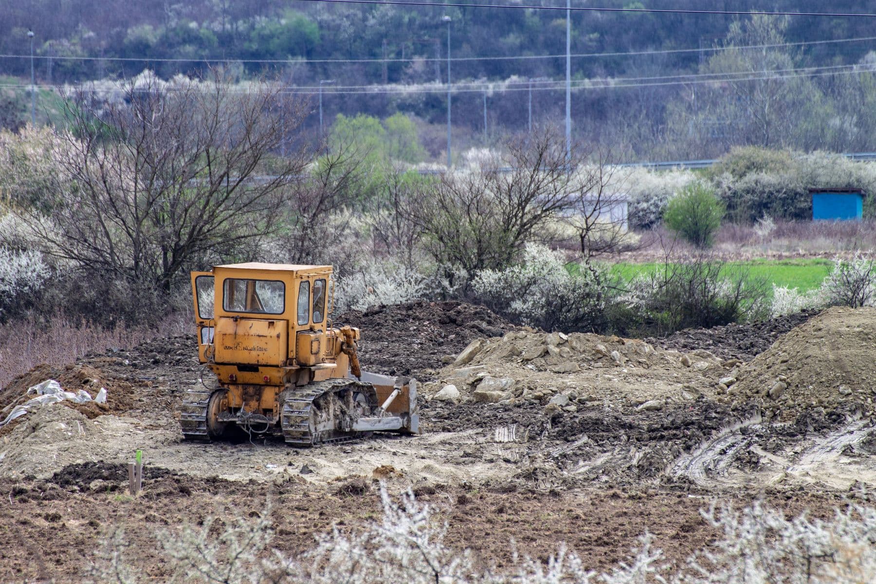 A yellow bulldozer on a cleared, bare-earth lot, with brush and bare trees around the edges.