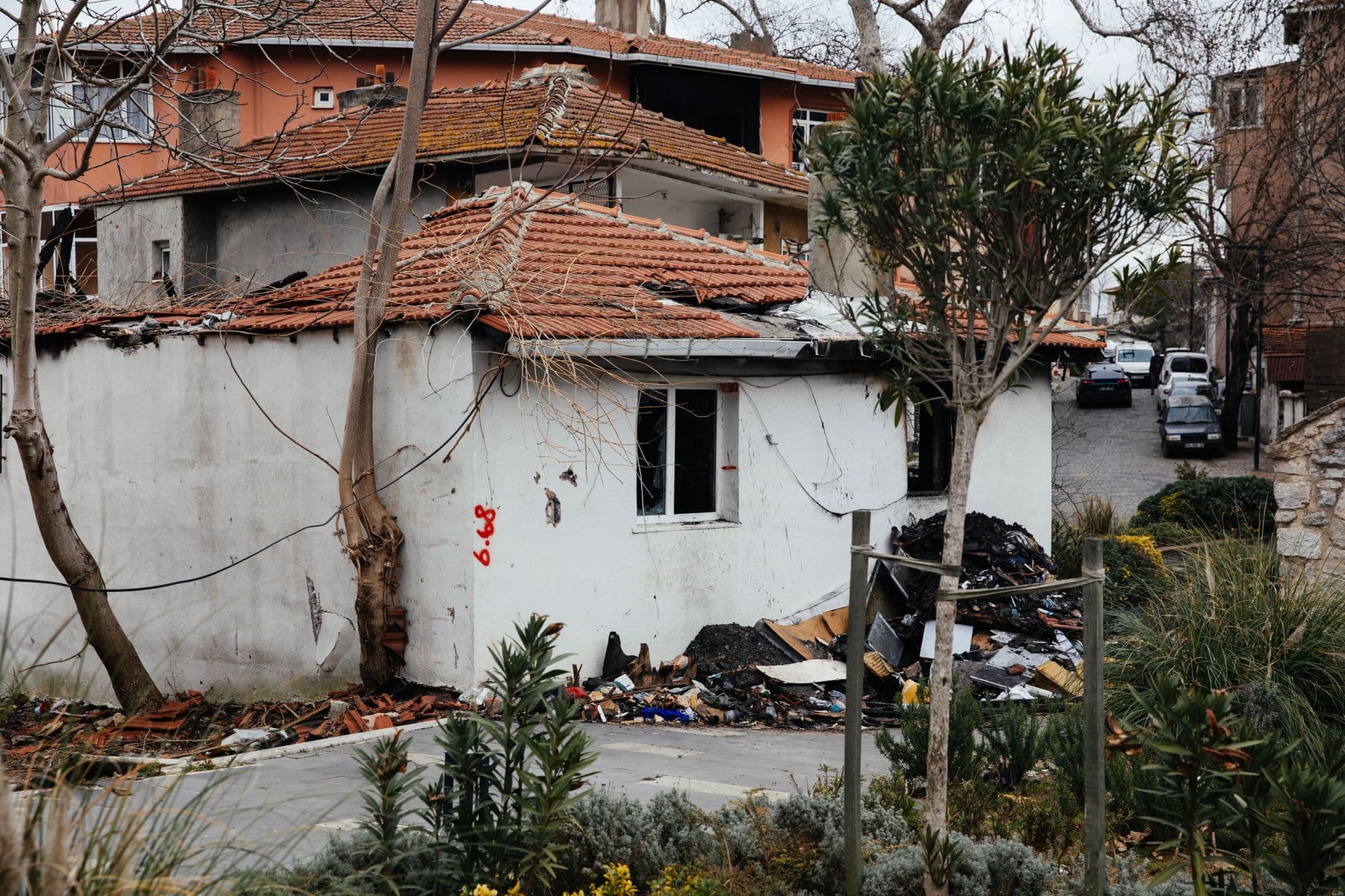 A small white single-story building with a broken tile roof, debris piled against it, on a street lined with trees.