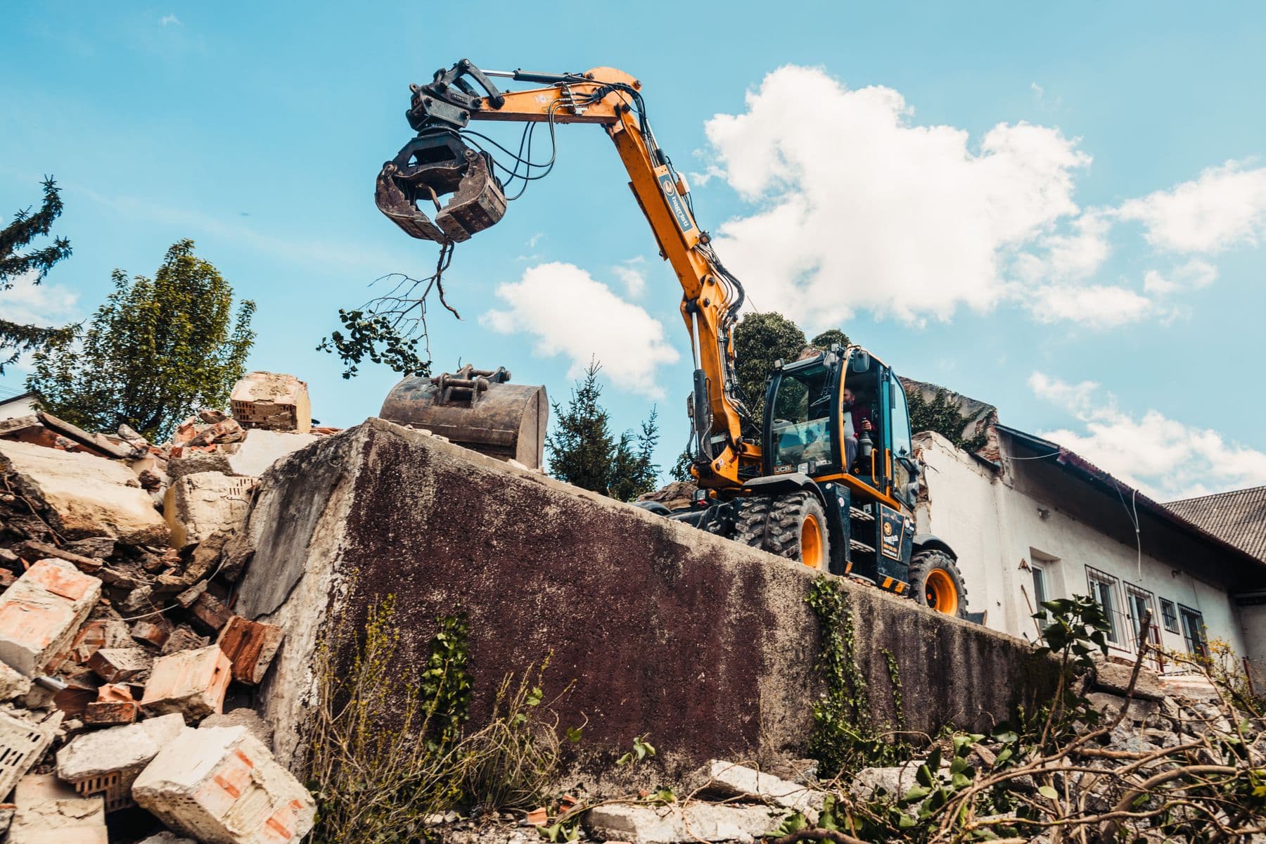 A wheeled excavator with a grab attachment beside a broken concrete wall and a pile of bricks, next to a low white building.