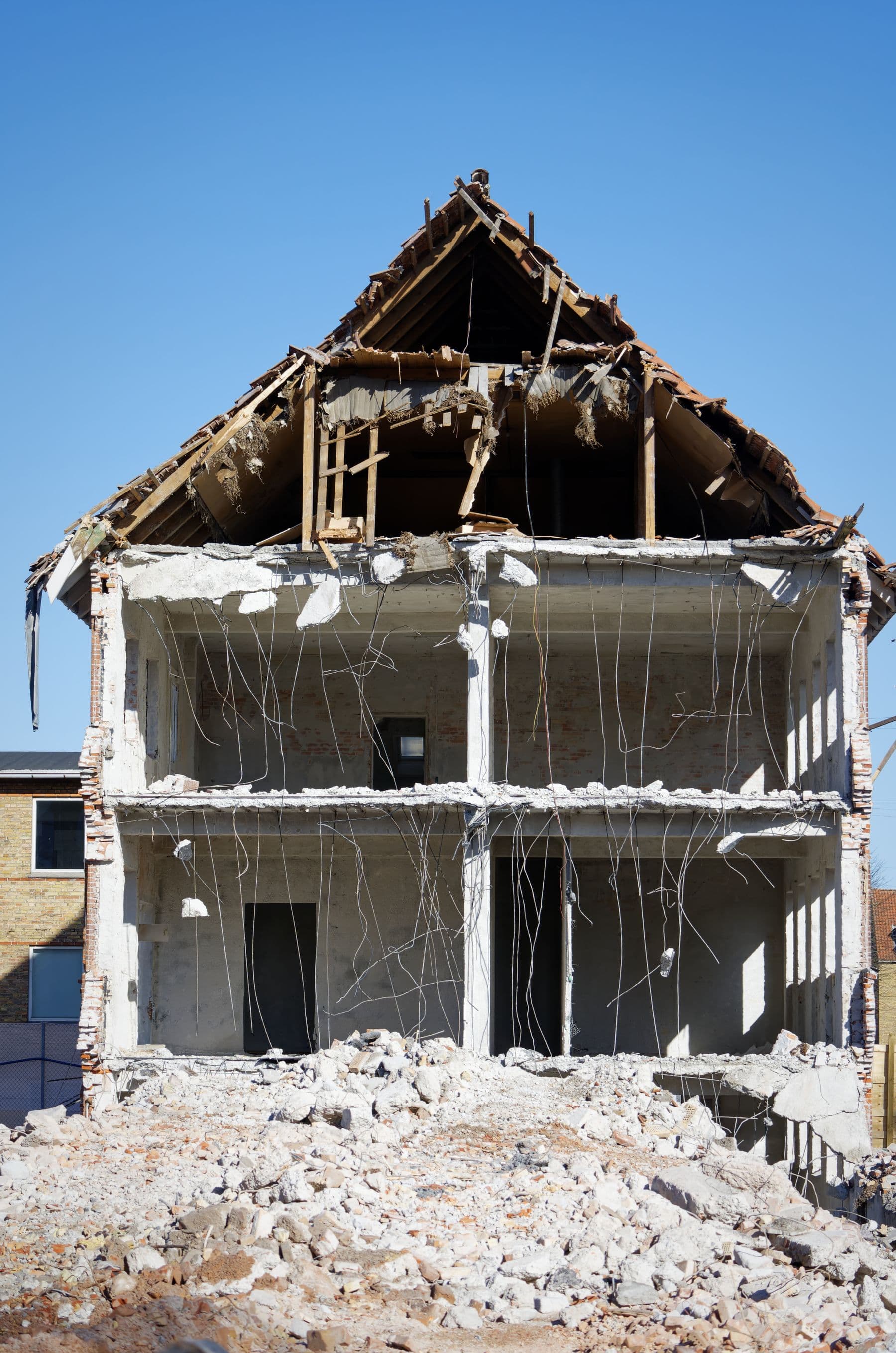 A two-story house with its outer walls removed, showing the concrete floor slabs, columns and the timber roof frame above a pile of rubble.