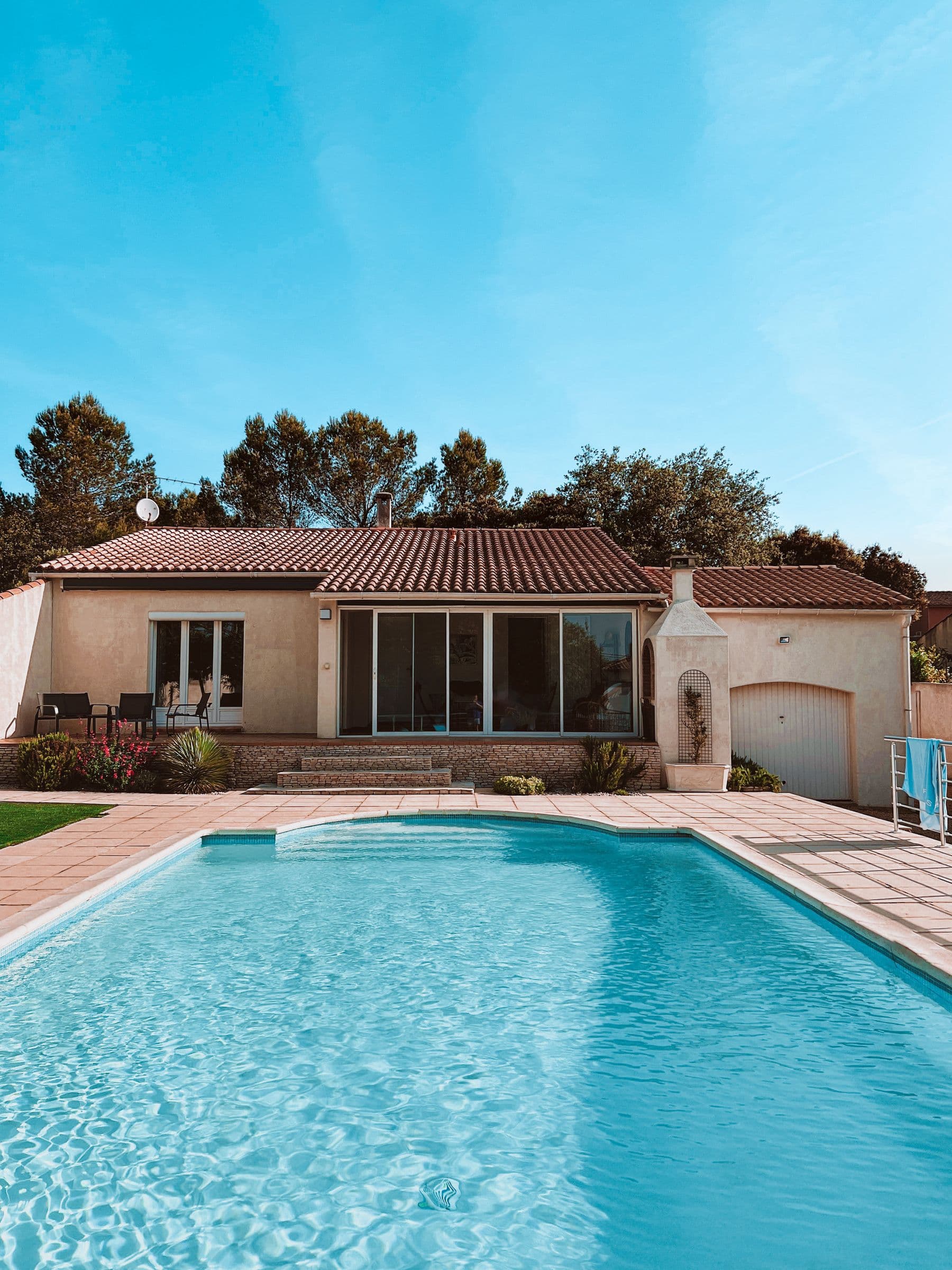 A rectangular in-ground swimming pool with a paved deck in front of a single-story house with a tiled roof.