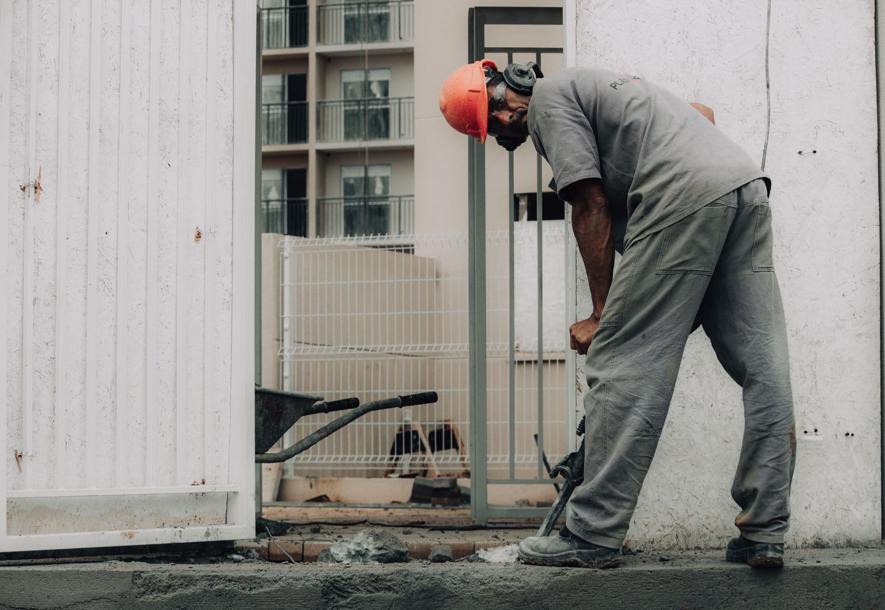 A worker in an orange hard hat bending over a jackhammer as it breaks up concrete outside a building entrance.