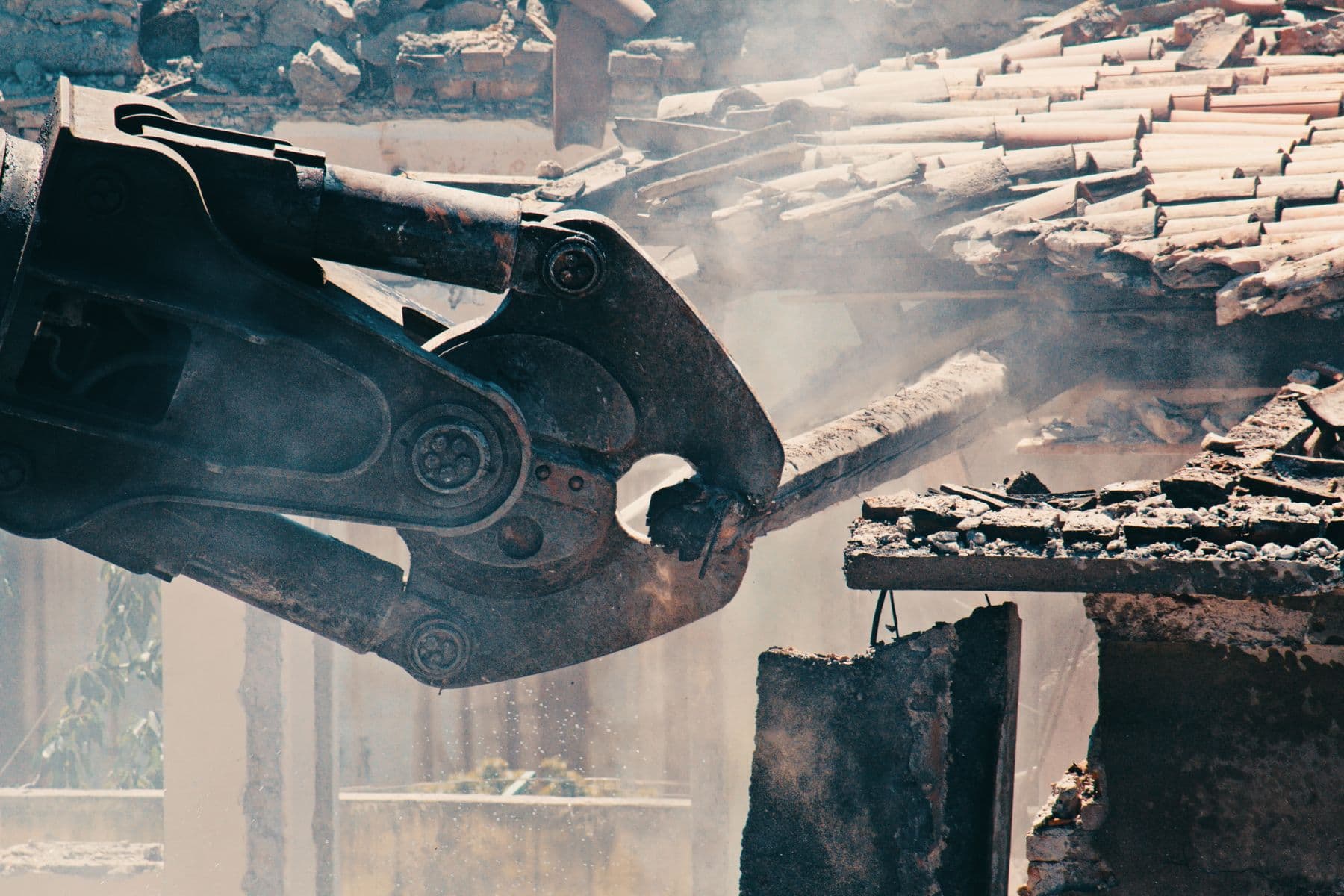 The steel jaws of a demolition shear biting into a clay-tile roof, with dust rising around it.