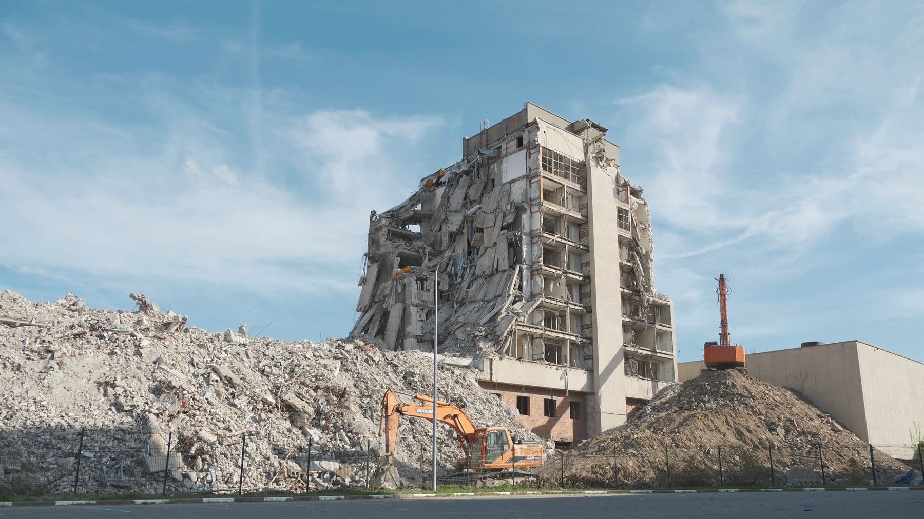 A partly demolished concrete apartment tower standing behind a long ridge of crushed rubble, with a small excavator at its base.