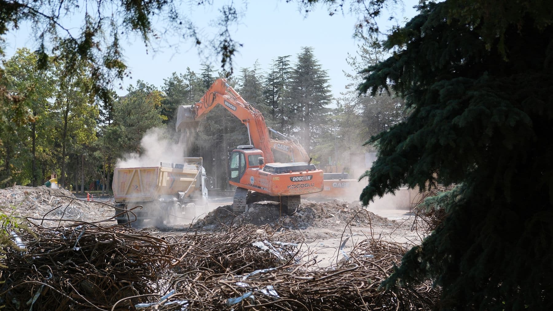 An orange excavator loading demolition debris into a dump truck in a cloud of dust, framed by pine trees.